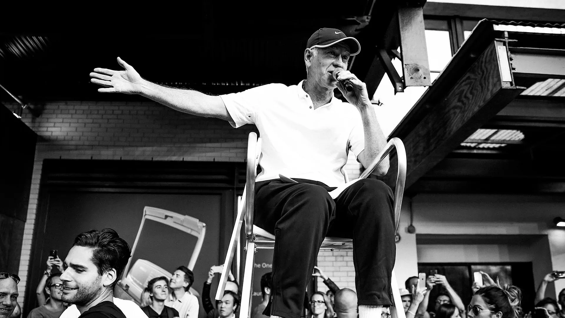 A man sits on a high chair, holding a microphone and gesturing with his arm outstretched, addressing a crowd of people below him at an indoor event. The image is in black and white.