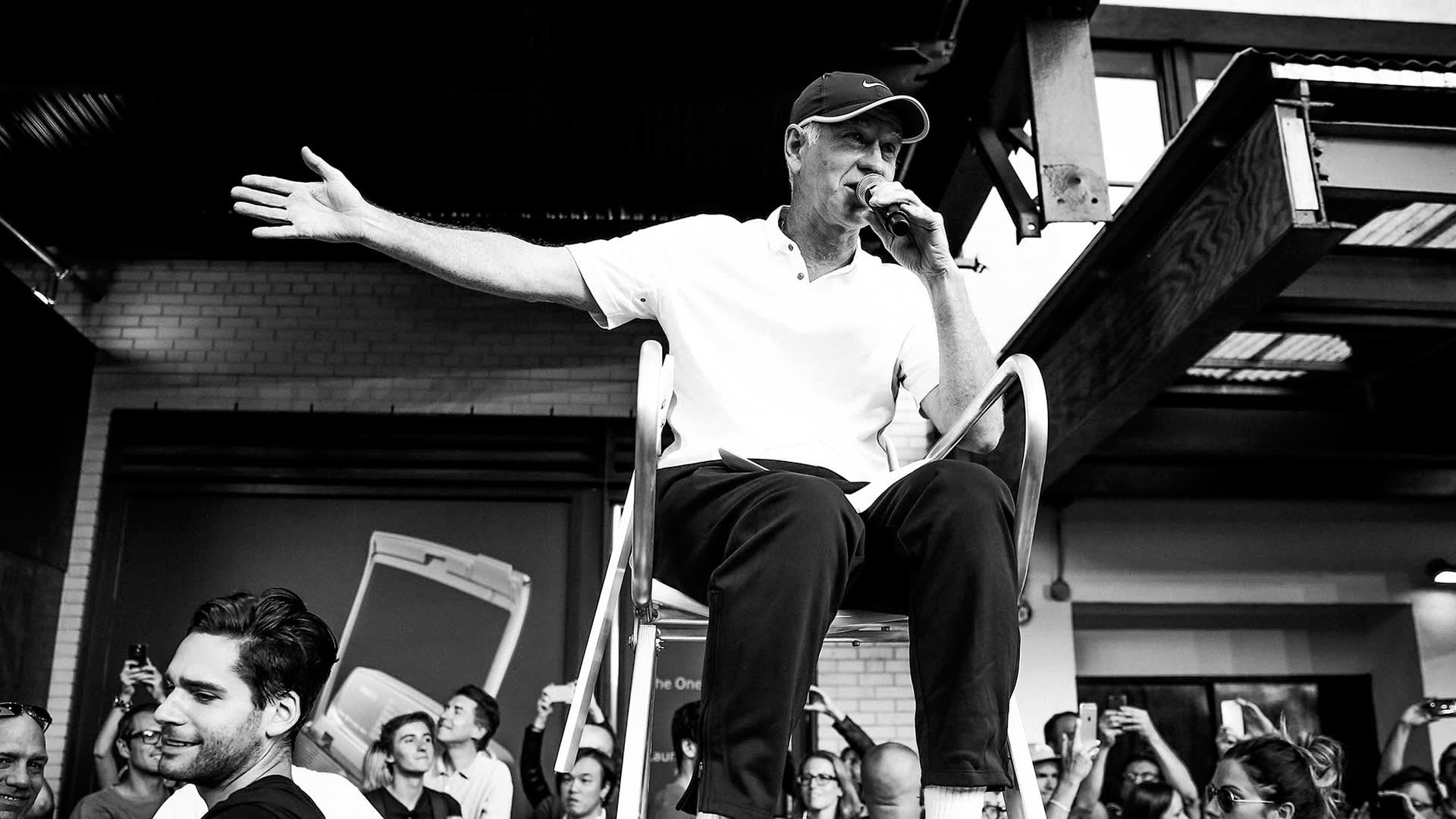 A man sits on a high chair, holding a microphone and gesturing with his arm outstretched, addressing a crowd of people below him at an indoor event. The image is in black and white.