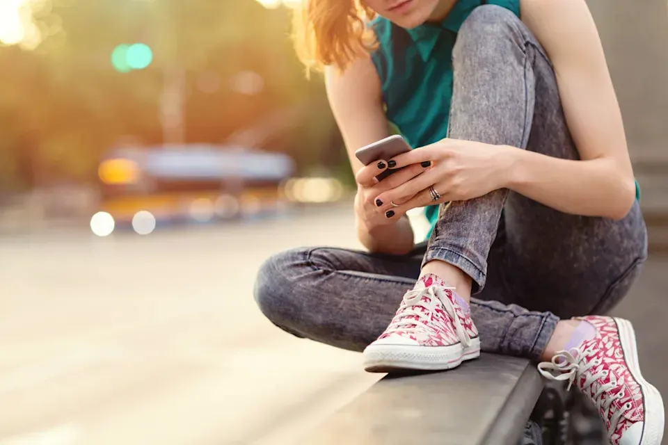 A young person sits outdoors on a ledge, wearing patterned pink sneakers, jeans, and a sleeveless shirt, looking down and using a smartphone. The background is blurred with sunlight and soft city lights.