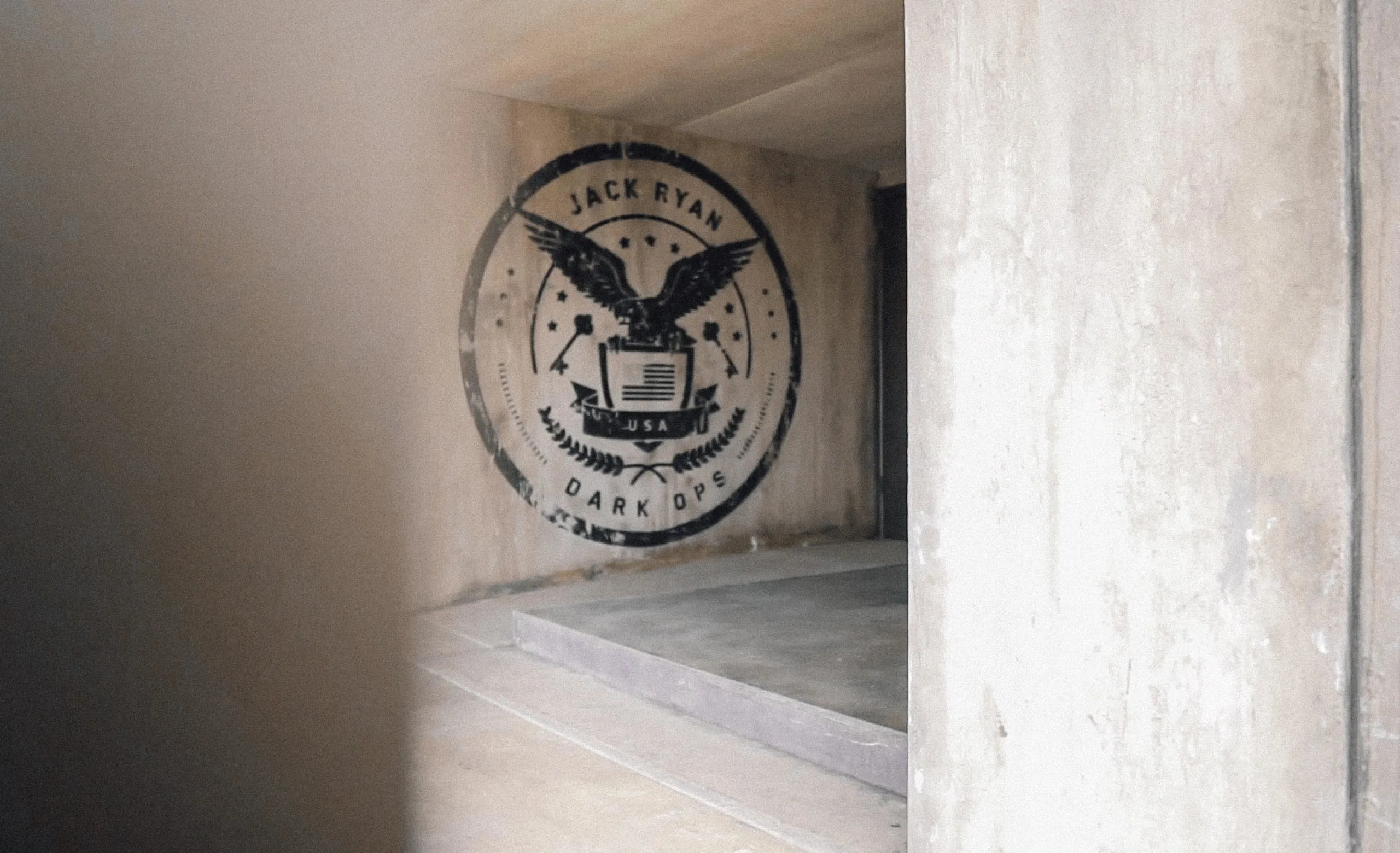 A concrete wall displays a circular emblem featuring an eagle holding arrows and an olive branch, with the text JACK RYAN DARK OPS and USA. The scene is indoors, with muted lighting and plain walls.