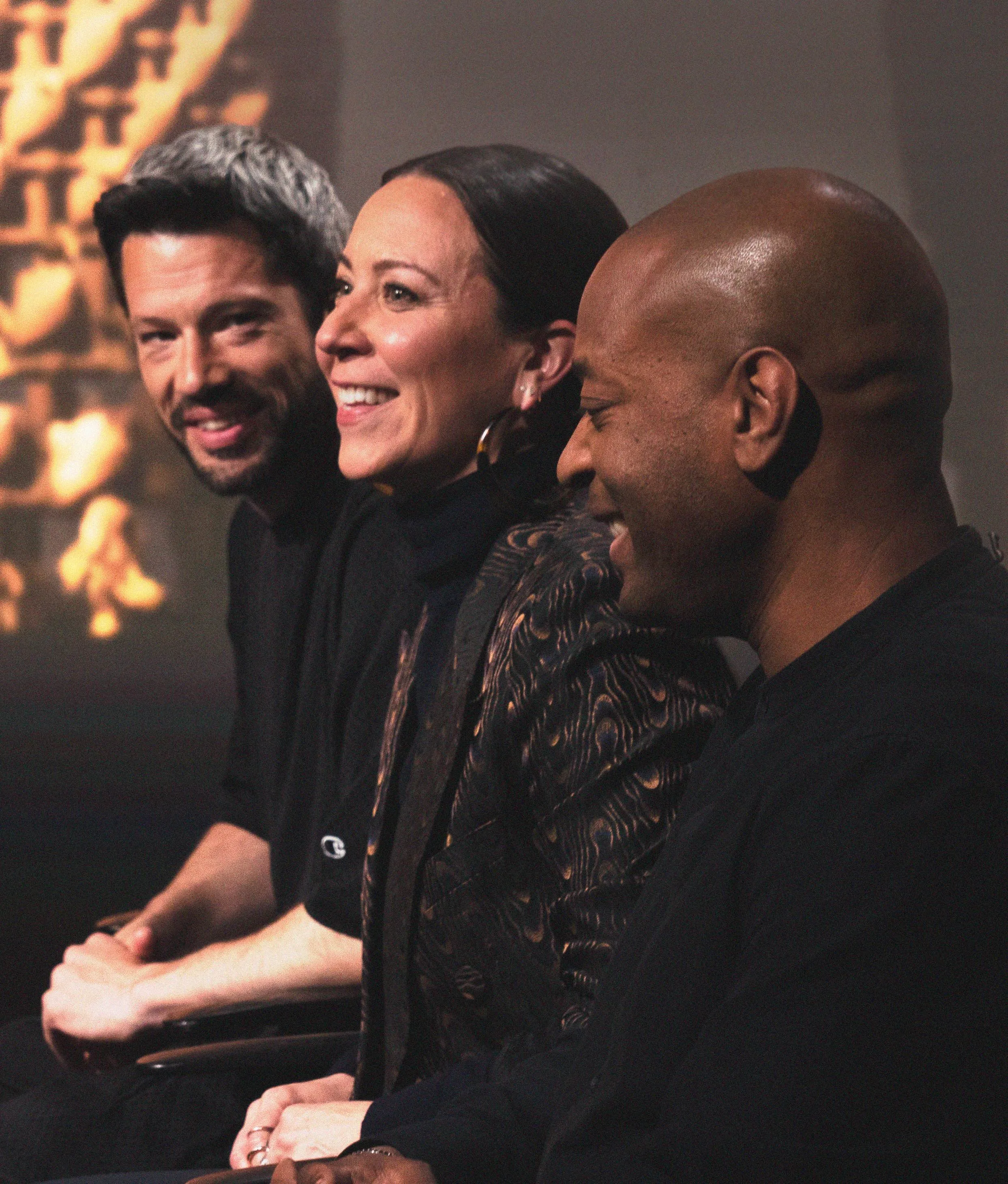 Three people dressed in dark clothing sit side by side, smiling and laughing, in a warmly lit setting with a blurred background.