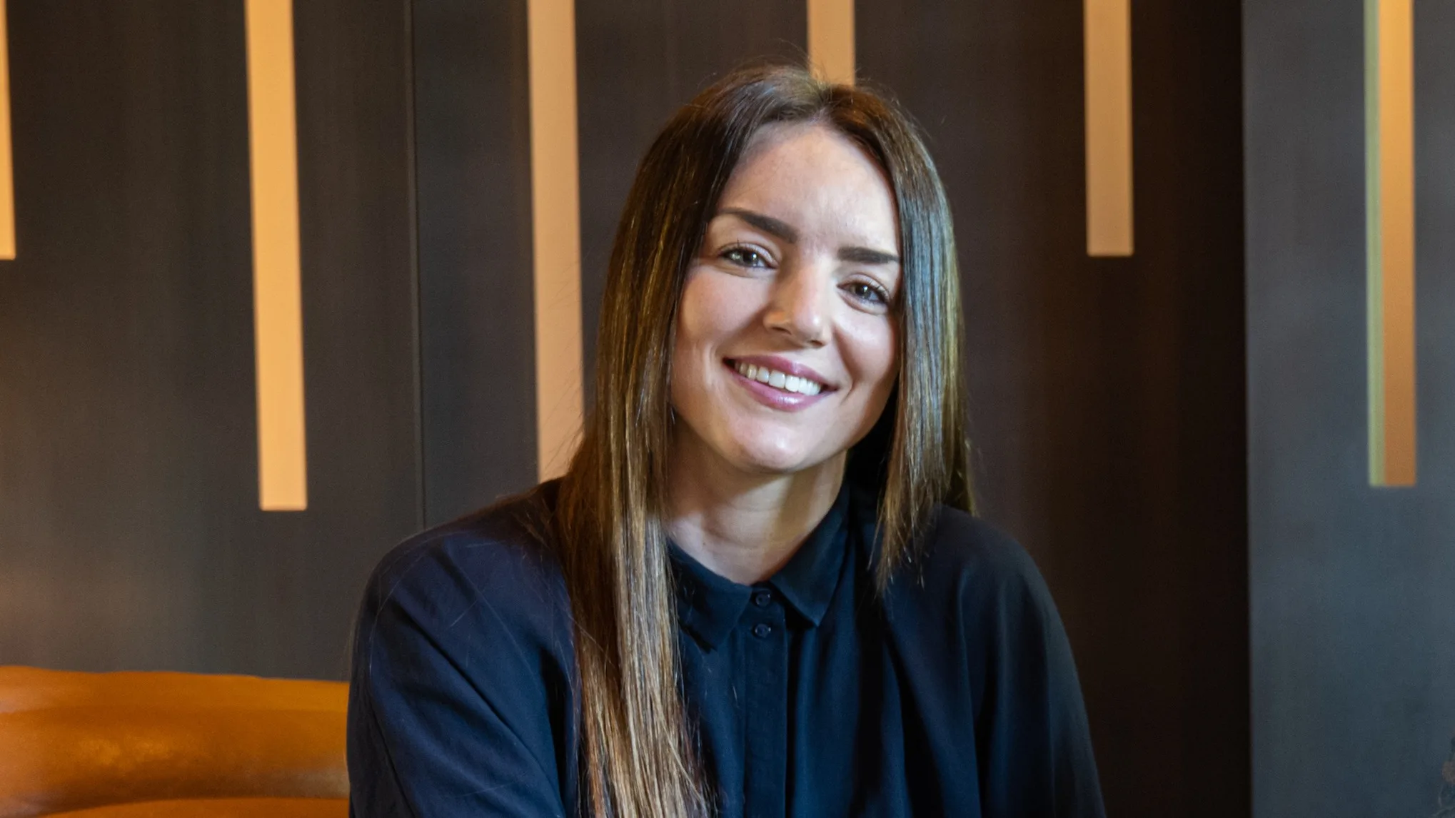 A woman with long brown hair, wearing a dark button-up shirt, smiles while sitting indoors against a modern background with vertical light strips.