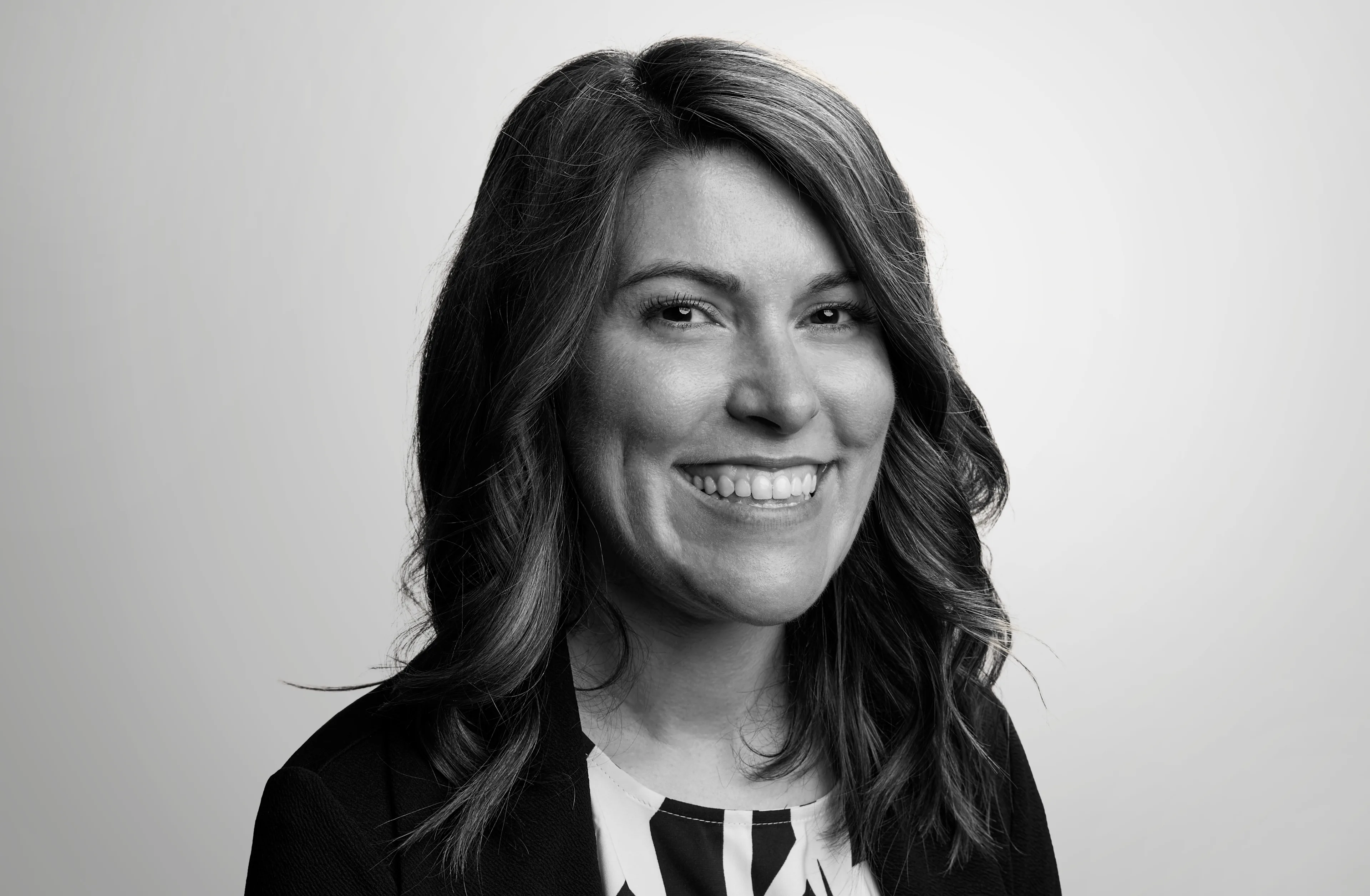 A woman with wavy, shoulder-length hair smiles at the camera. She is wearing a dark blazer over a patterned blouse. The background is a light, plain gradient.
