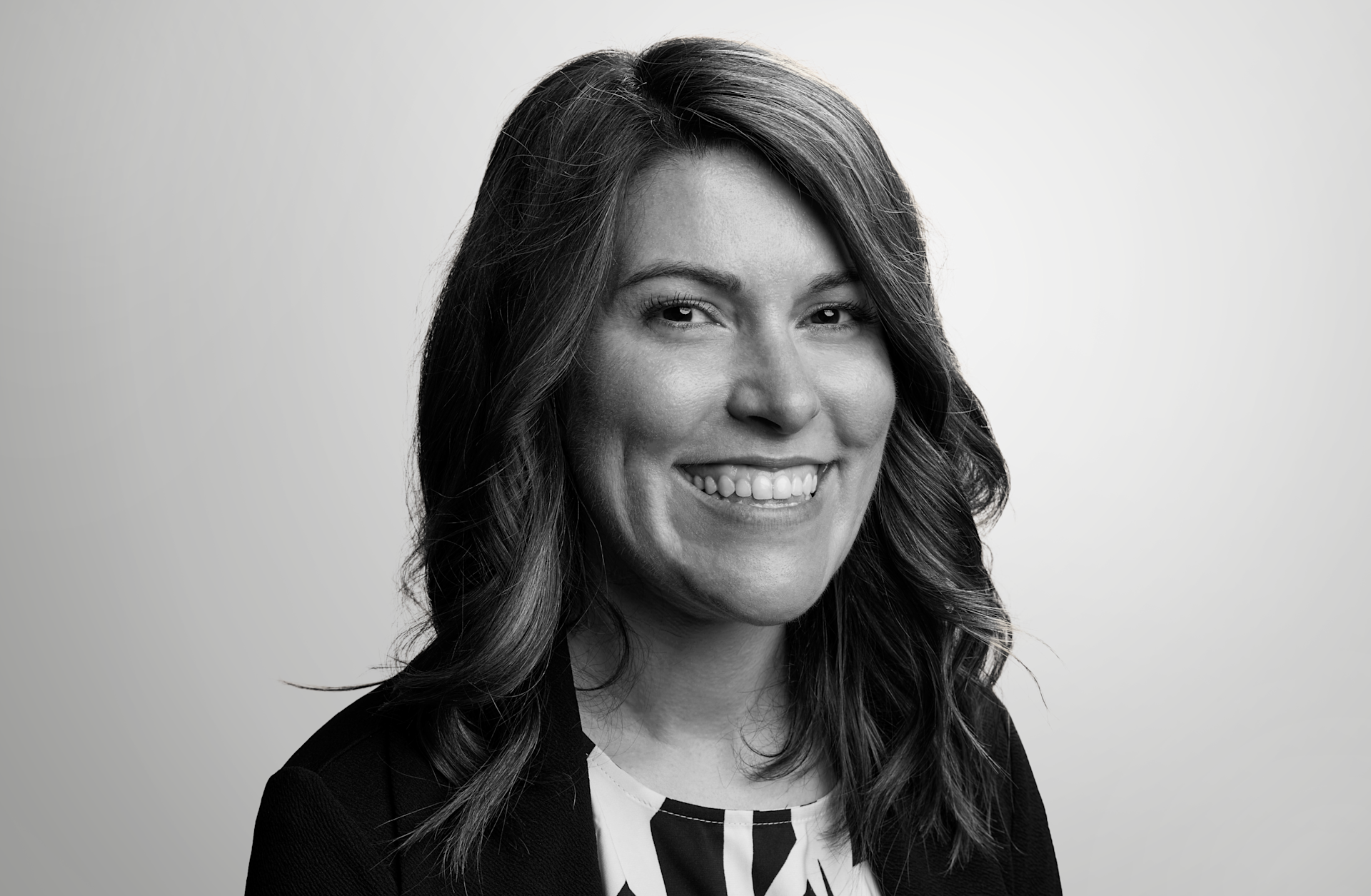 A woman with wavy, shoulder-length hair smiles at the camera. She is wearing a dark blazer over a patterned blouse. The background is a light, plain gradient.