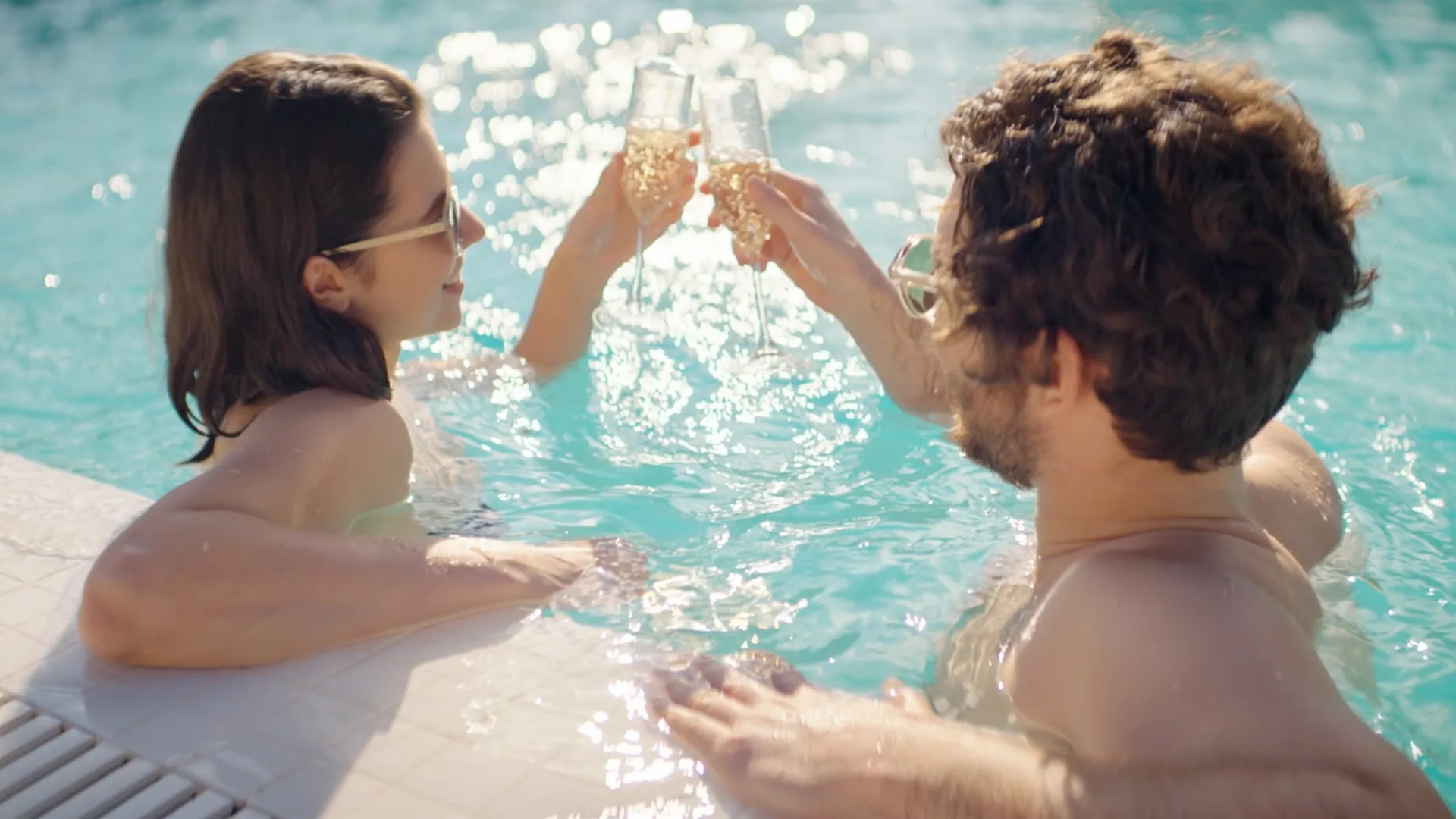 Two people relaxing in a swimming pool clink champagne glasses together, enjoying a sunny day. They are wearing sunglasses and appear to be celebrating or toasting by the poolside.