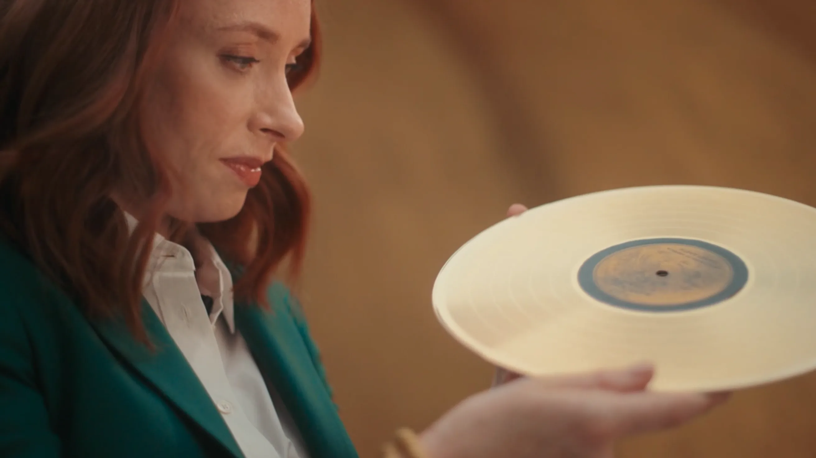 A woman with reddish-brown hair, wearing a green blazer and white shirt, looks thoughtfully at a gold-colored vinyl record that she is holding in her hands.