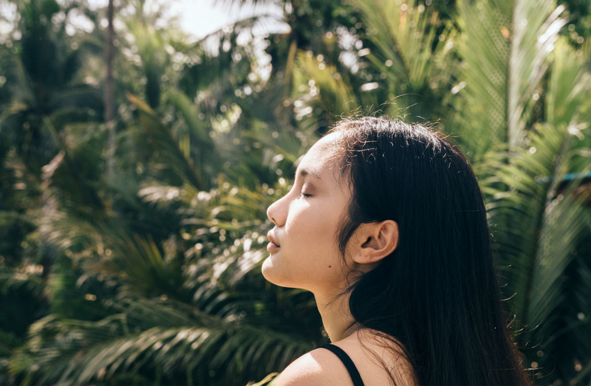 A woman with long dark hair stands outdoors with her eyes closed, enjoying the sunlight. Lush green palm trees and foliage surround her in the background.