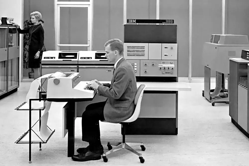 A man sits at a desk typing on an early computer terminal, with large IBM mainframe computers and equipment in the background. A woman stands near additional machines in the room. The scene appears from the mid-20th century.