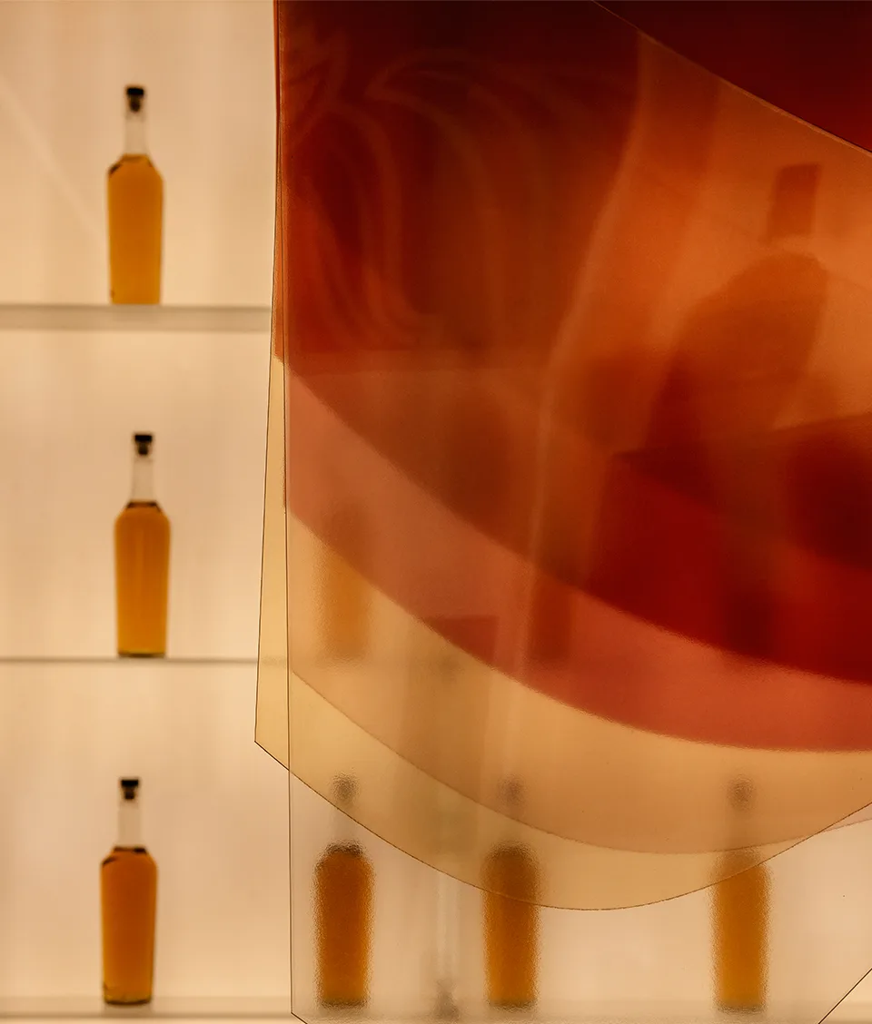 Three shelves each hold two bottles of amber liquid against a softly lit background, with a sheer, reddish curtain draped partly in front, creating layered shadows and a warm, abstract effect.