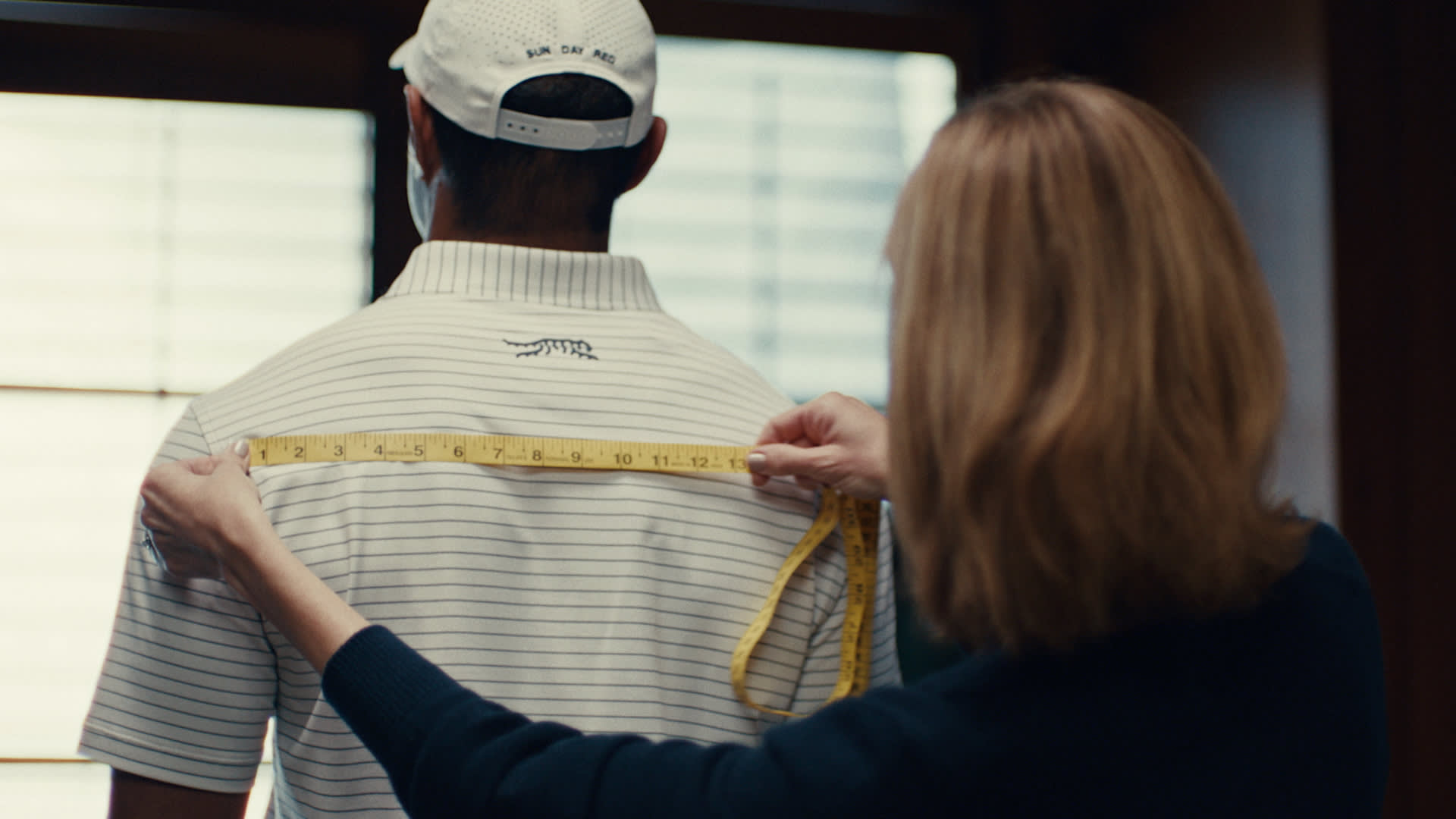 A person measures the back width of another person, who is wearing a striped polo shirt and a cap, using a yellow measuring tape indoors in front of a window.