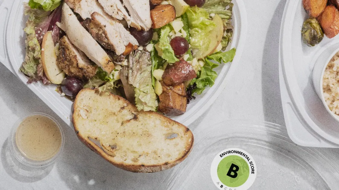 A takeout container with salad topped with sliced grilled chicken, grapes, and roasted sweet potatoes, served with a slice of bread and a small cup of dressing. A container lid labeled with a B environmental score is in view.