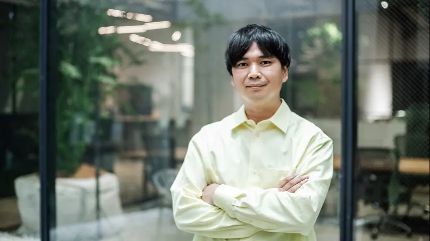 A man with short dark hair stands indoors with his arms crossed, wearing a light yellow shirt, in front of glass walls of an office with plants and blurred furniture in the background.