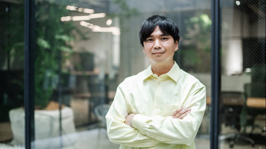 A man with short dark hair stands indoors with his arms crossed, wearing a light yellow shirt, in front of glass walls of an office with plants and blurred furniture in the background.