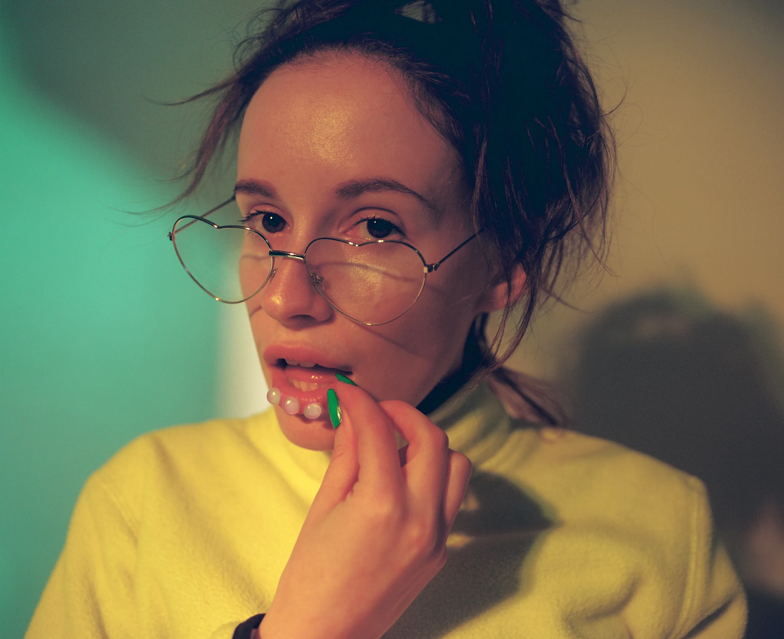 A person with tousled hair and round glasses wears a yellow turtleneck and holds a string of pearls to their lips, looking directly at the camera with a neutral expression under soft, moody lighting.