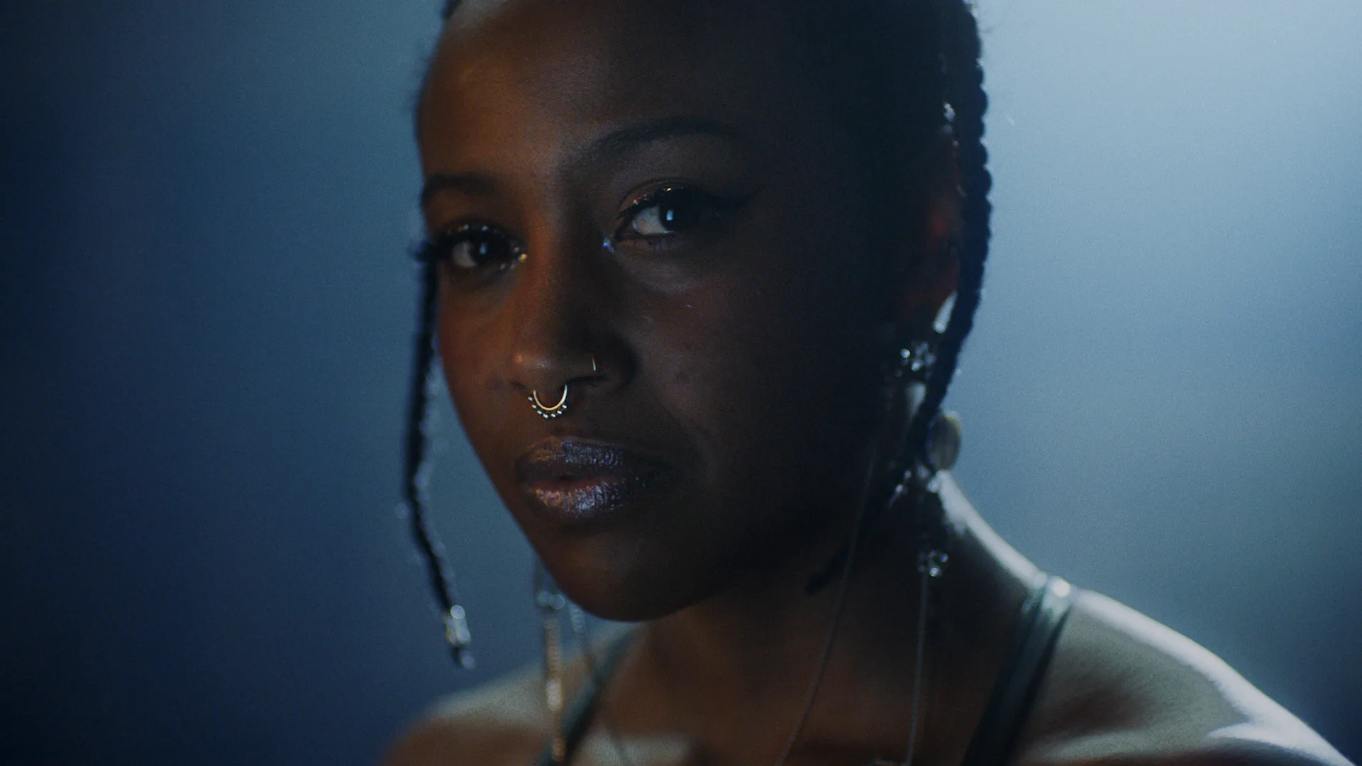 A person with braided hair, a septum piercing, and hoop earrings looks directly at the camera. The lighting is soft and moody, creating a gentle highlight on their face against a dark background.