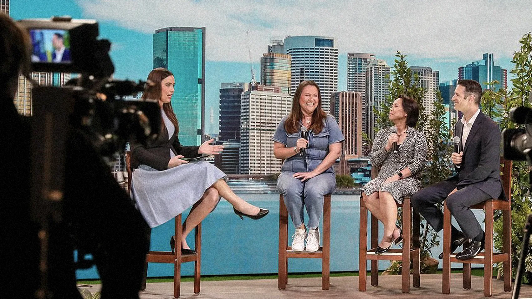Four people sit on high stools, smiling and talking during a panel discussion, with a city skyline backdrop behind them. A camera operator films the scene from the left foreground.