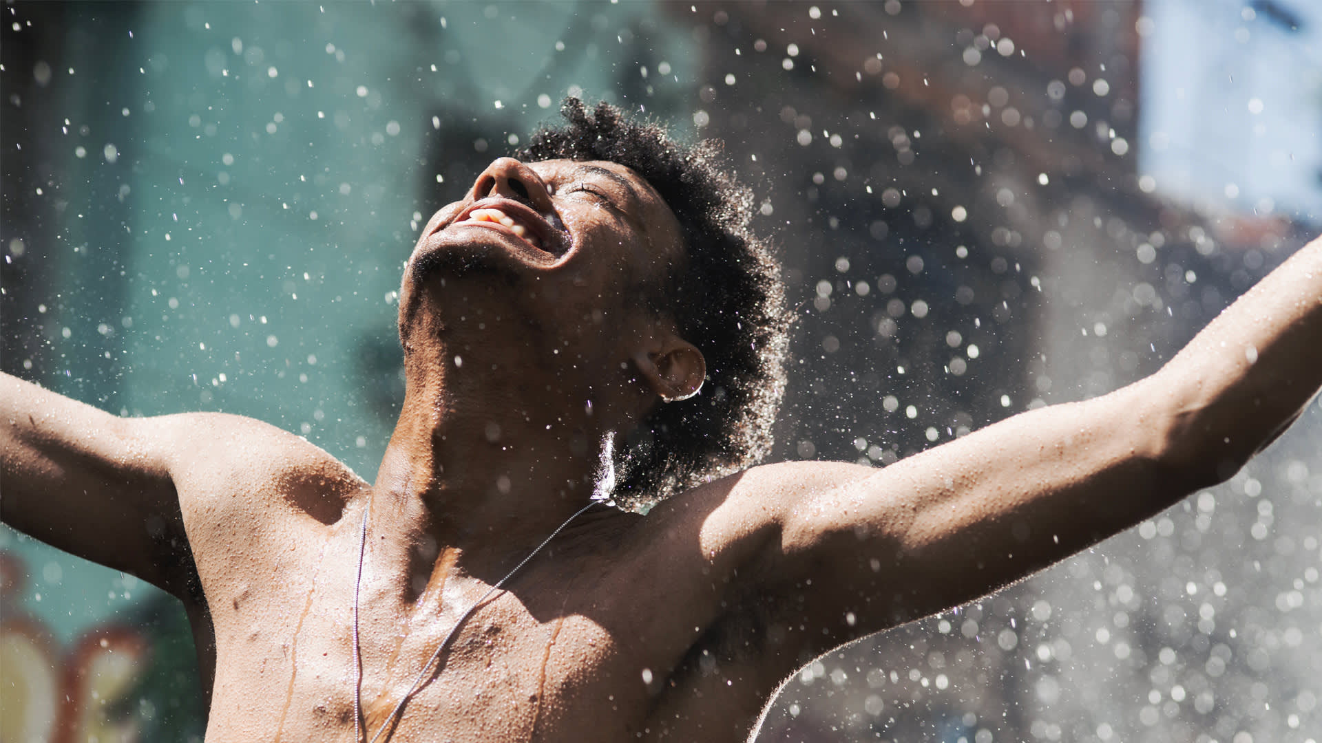A person smiles joyfully with eyes closed and arms outstretched, standing in sunlight with water droplets falling around them.