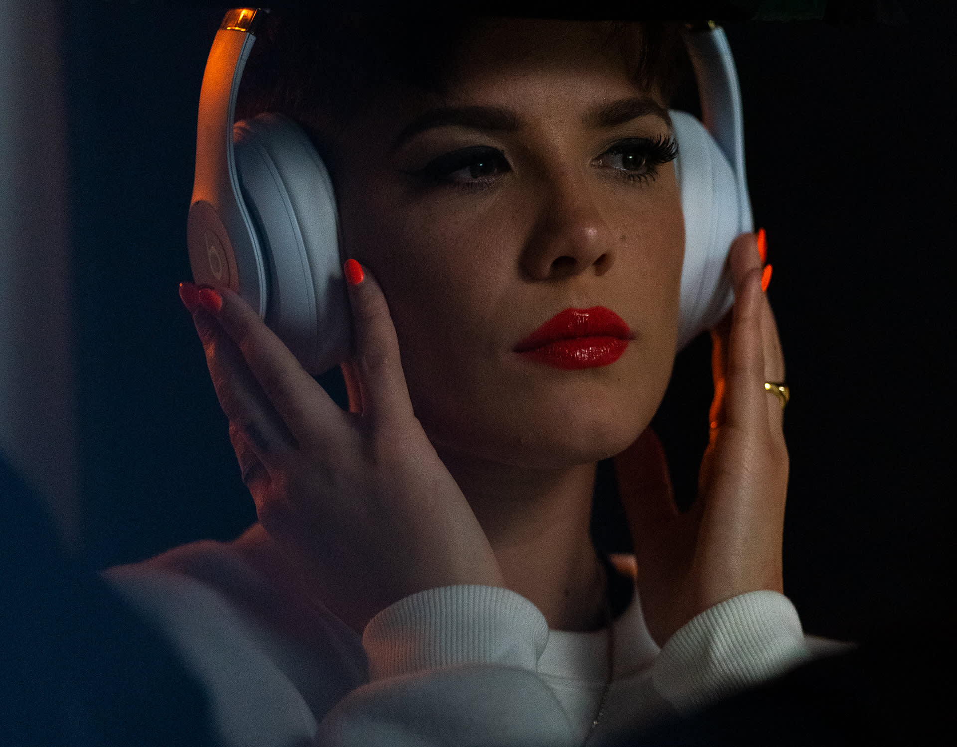 A person with red lipstick and nail polish wears white headphones, holding them with both hands. They have short hair, winged eyeliner, and a focused expression in dim lighting.