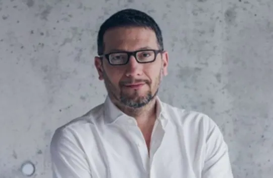 A man with short dark hair and a beard, wearing glasses and a white shirt, stands in front of a gray, textured concrete wall with a neutral expression.