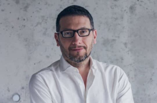 A man with short dark hair and a beard, wearing glasses and a white shirt, stands in front of a gray, textured concrete wall with a neutral expression.