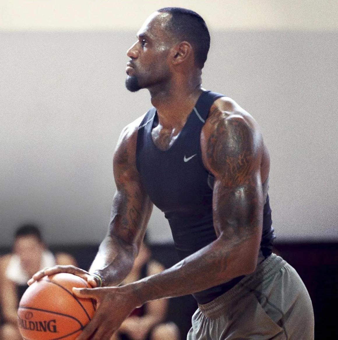 A muscular man wearing a black sleeveless athletic shirt holds a basketball, preparing to shoot. He stands in a gym with blurred people in the background and looks focused on the hoop.