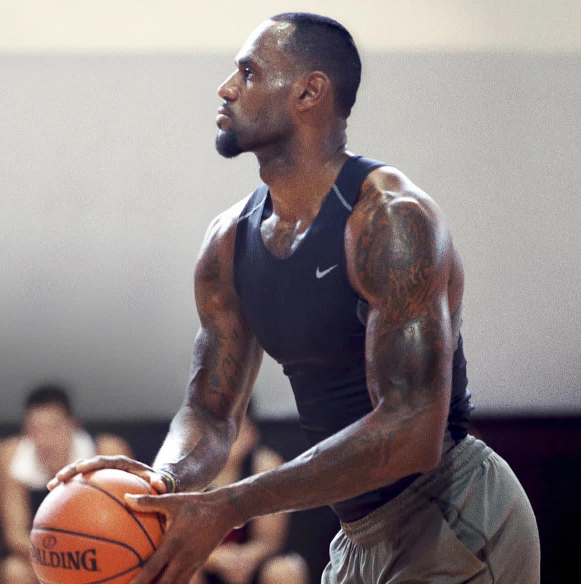 A muscular man wearing a black sleeveless athletic shirt holds a basketball, preparing to shoot. He stands in a gym with blurred people in the background and looks focused on the hoop.