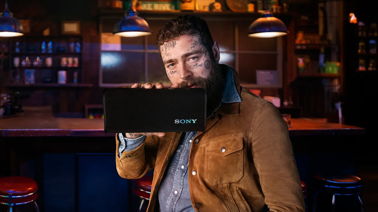 A bearded man with face tattoos holds a black Sony portable speaker in front of his mouth while sitting in a dimly lit bar with red stools and hanging lights.
