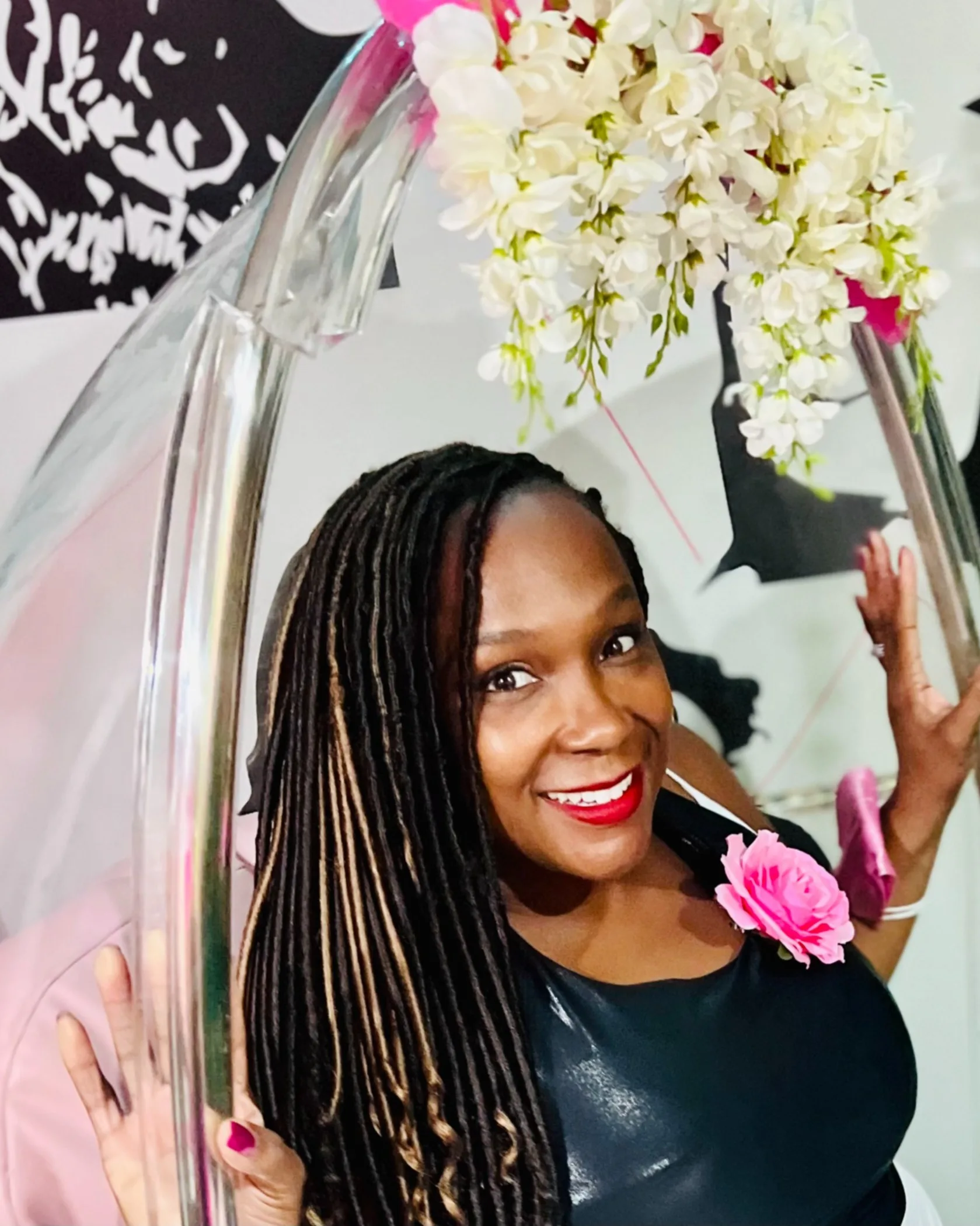 A smiling woman with long braids wearing a black top and pink flower accessory poses inside a clear hanging chair decorated with white flowers. Black and white art is visible in the background.