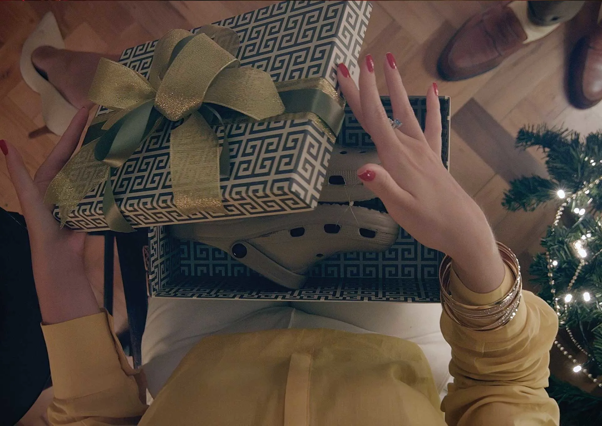 A person with red nail polish opens a decorative gift box with a gold ribbon, revealing a pair of Crocs inside. A Christmas tree with lights and presents is visible on the right, and two people stand nearby.