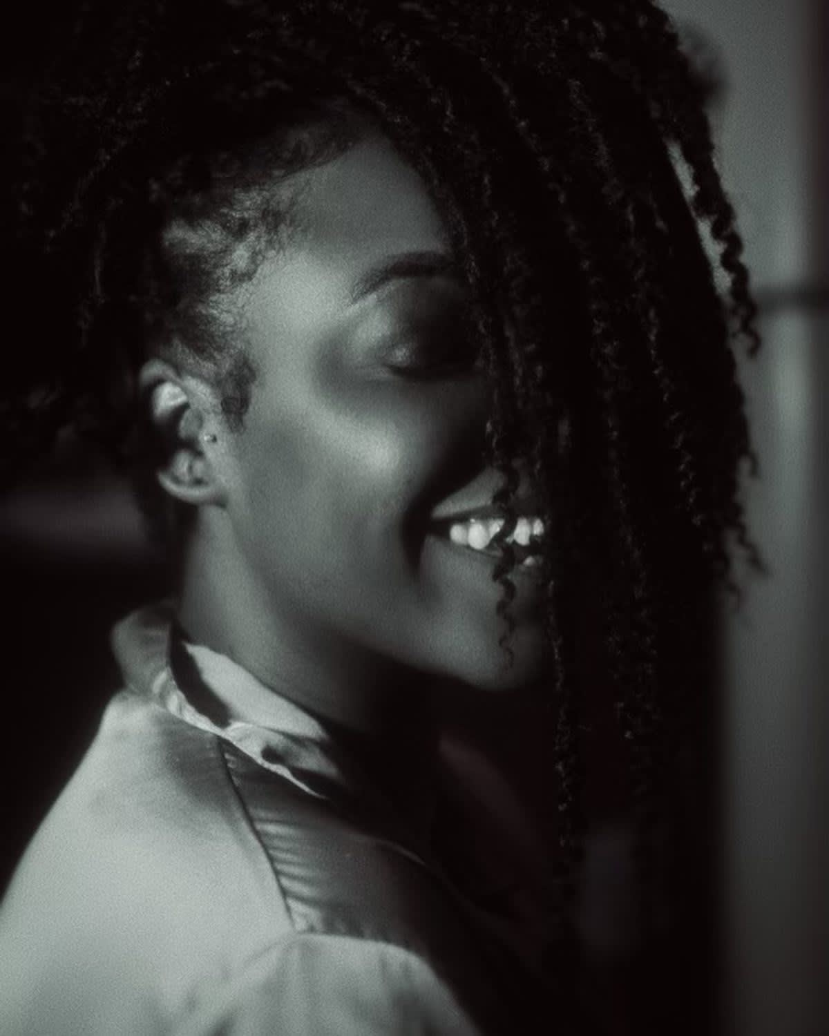 A woman with curly hair partially covering her face smiles warmly in a softly lit, black-and-white photo, wearing a satin robe and looking relaxed and happy.