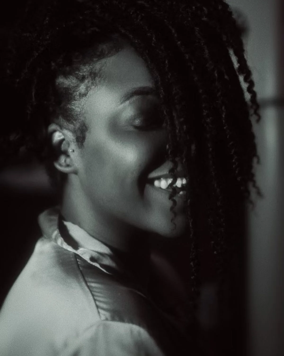 A woman with curly hair partially covering her face smiles warmly in a softly lit, black-and-white photo, wearing a satin robe and looking relaxed and happy.