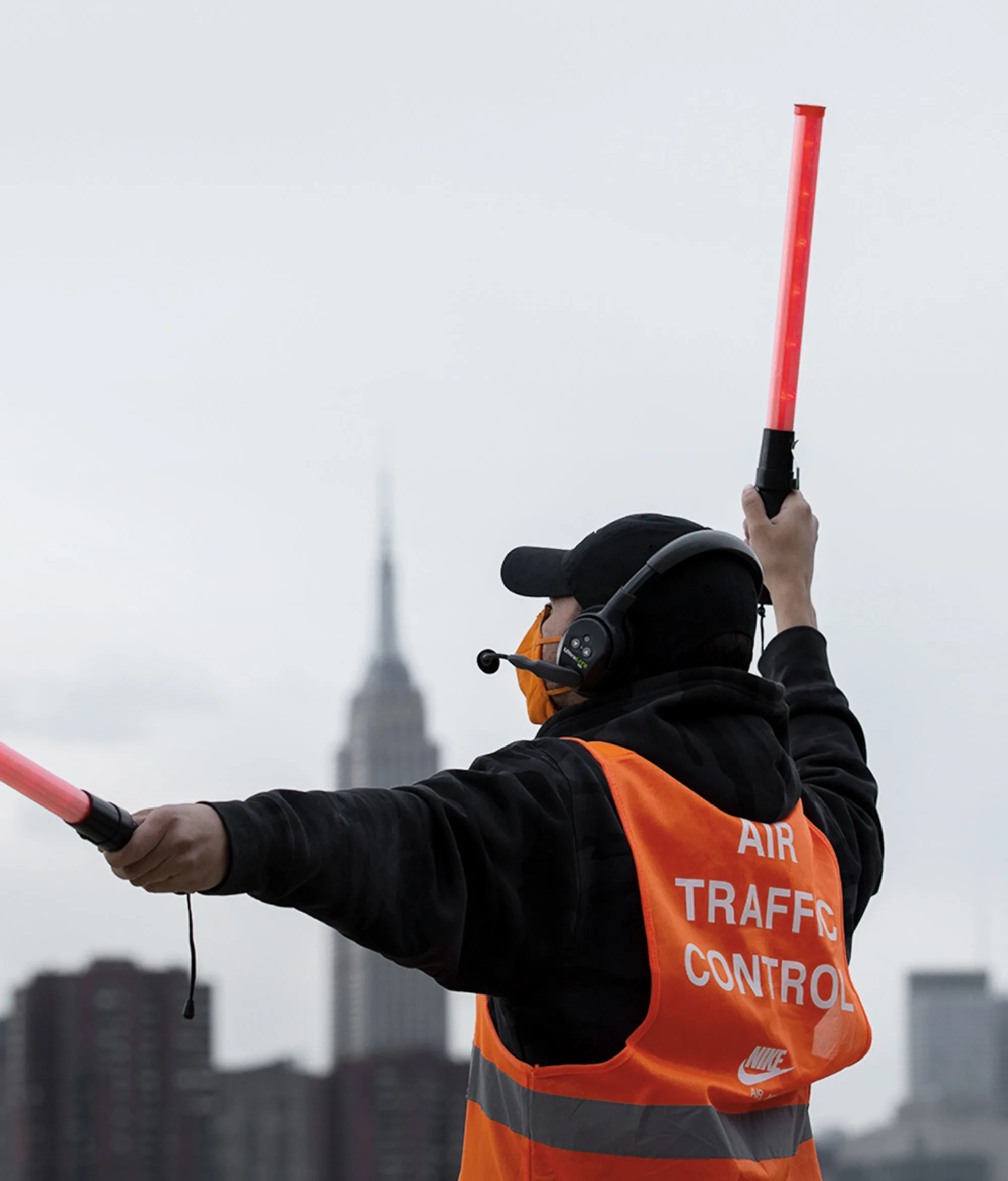 A person wearing an orange Air Traffic Control vest and headset directs air traffic with two red wands, with a city skyline and the Empire State Building visible in the background.