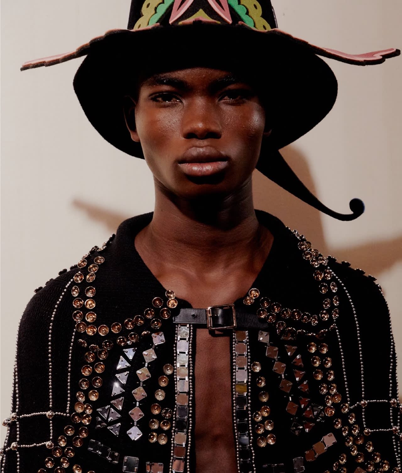 A person with dark skin wears a large, elaborate, black hat with colorful detailing and a wide brim, and a black jacket adorned with shiny metallic studs and square embellishments, set against a plain background.