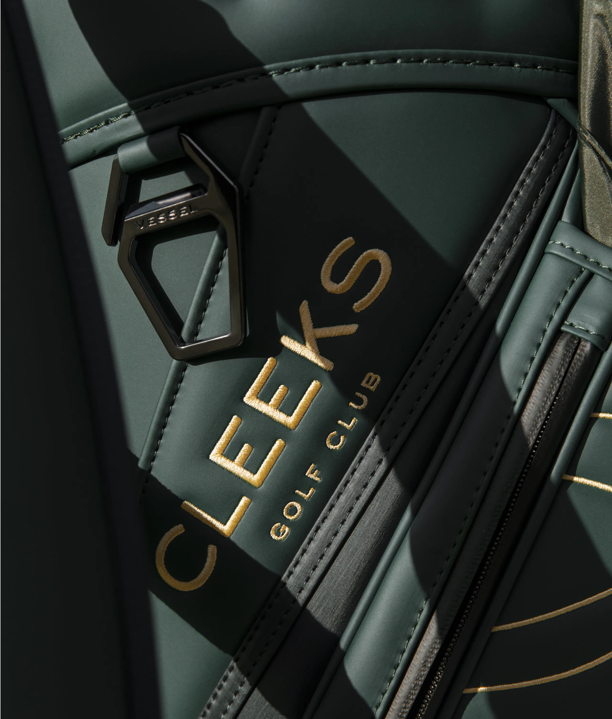 Close-up of a dark green golf bag with the words CLEEKS GOLF CLUB in gold lettering and a metal fastener, partially lit by sunlight.