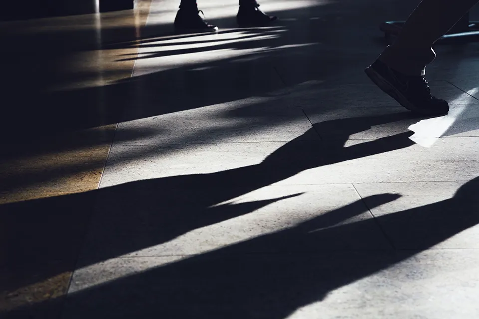 Close-up of peoples legs and feet walking indoors, casting long shadows on a sunlit tiled floor.