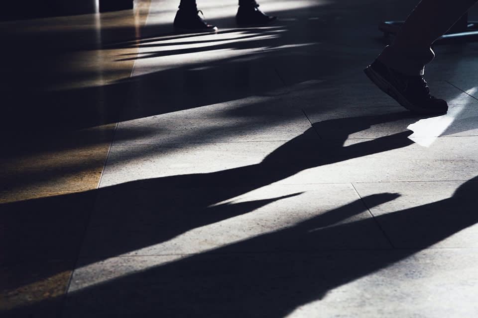 Close-up of peoples legs and feet walking indoors, casting long shadows on a sunlit tiled floor.