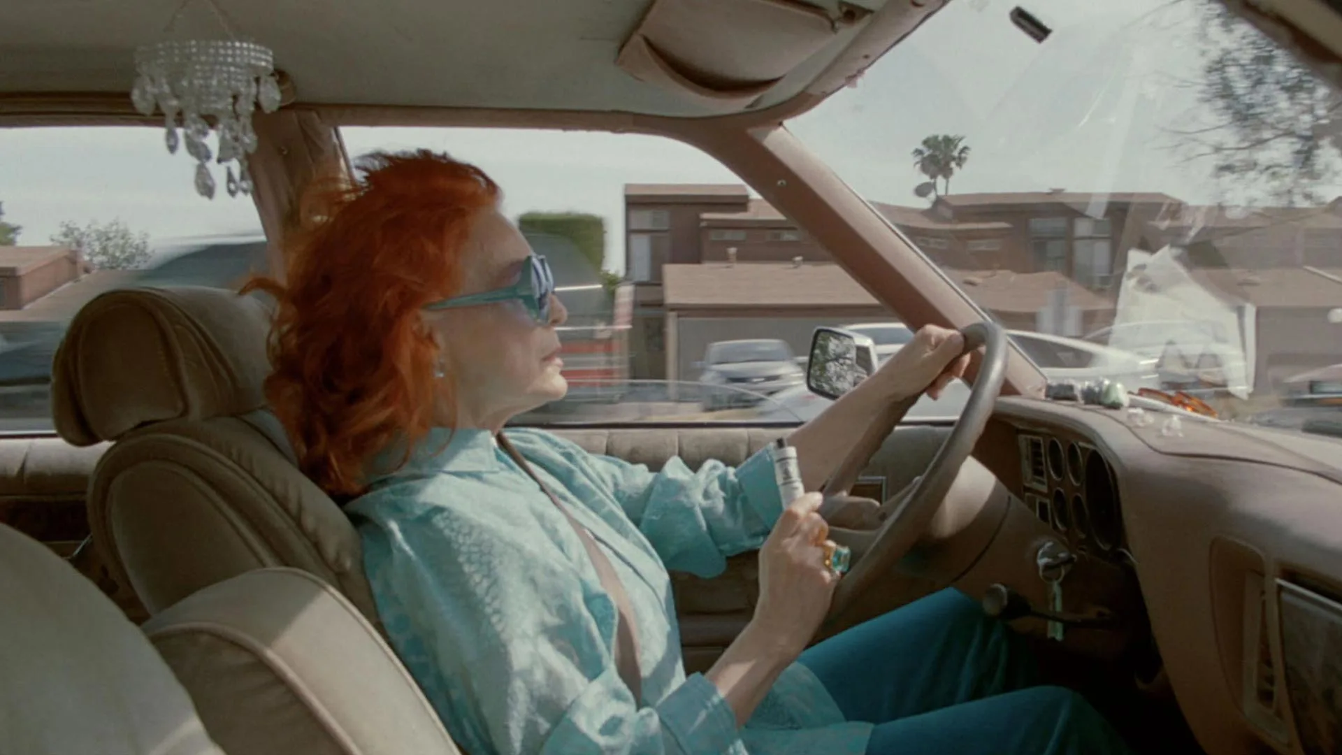 A woman with red hair and blue sunglasses drives a car through a suburban neighborhood during the day, holding a small object in her left hand and wearing a light blue outfit.