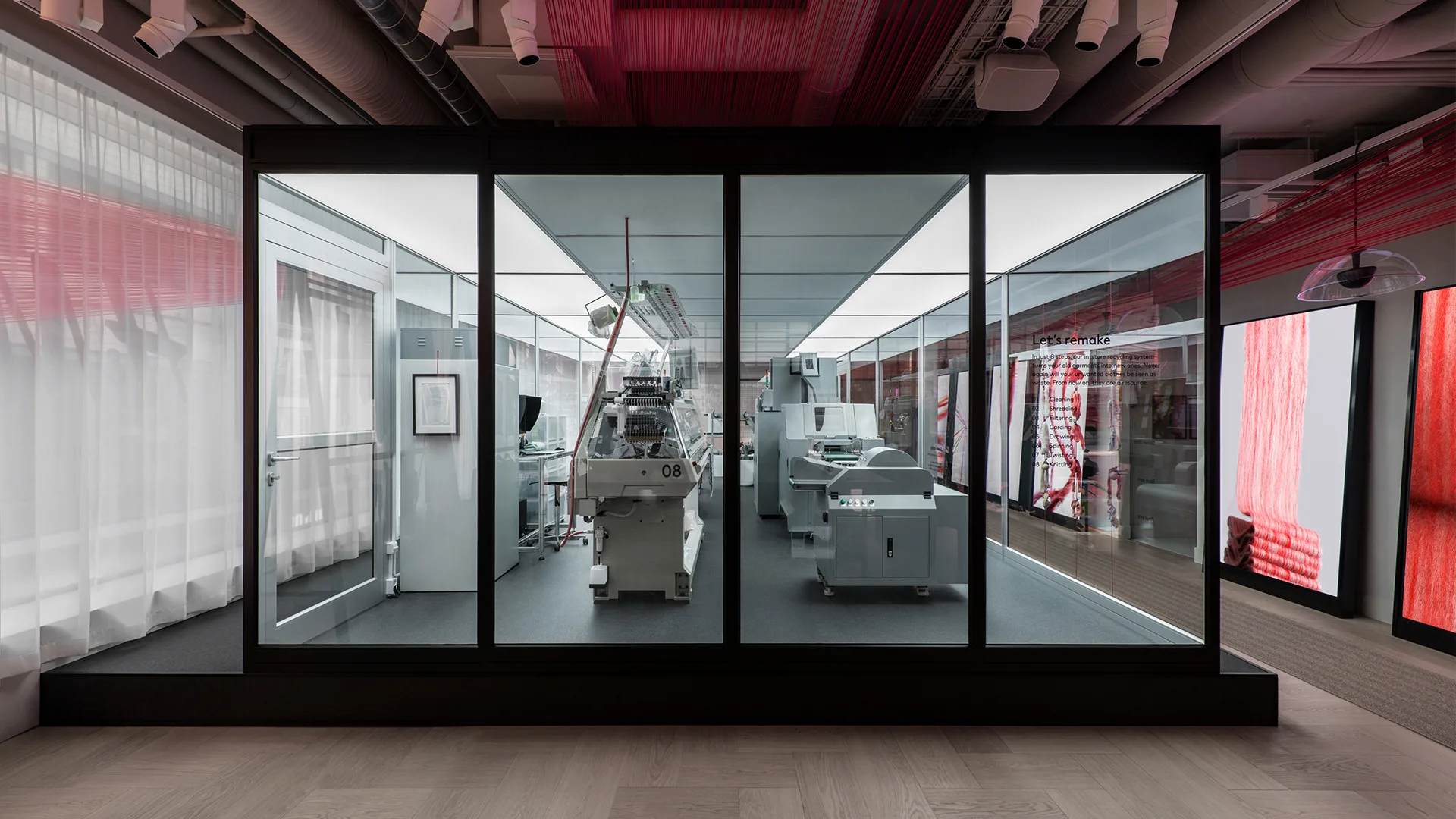 A modern exhibit displays automated textile machines behind glass panels in a well-lit showroom, with red and white fabric accents and exposed ceiling pipes visible in the surroundings.