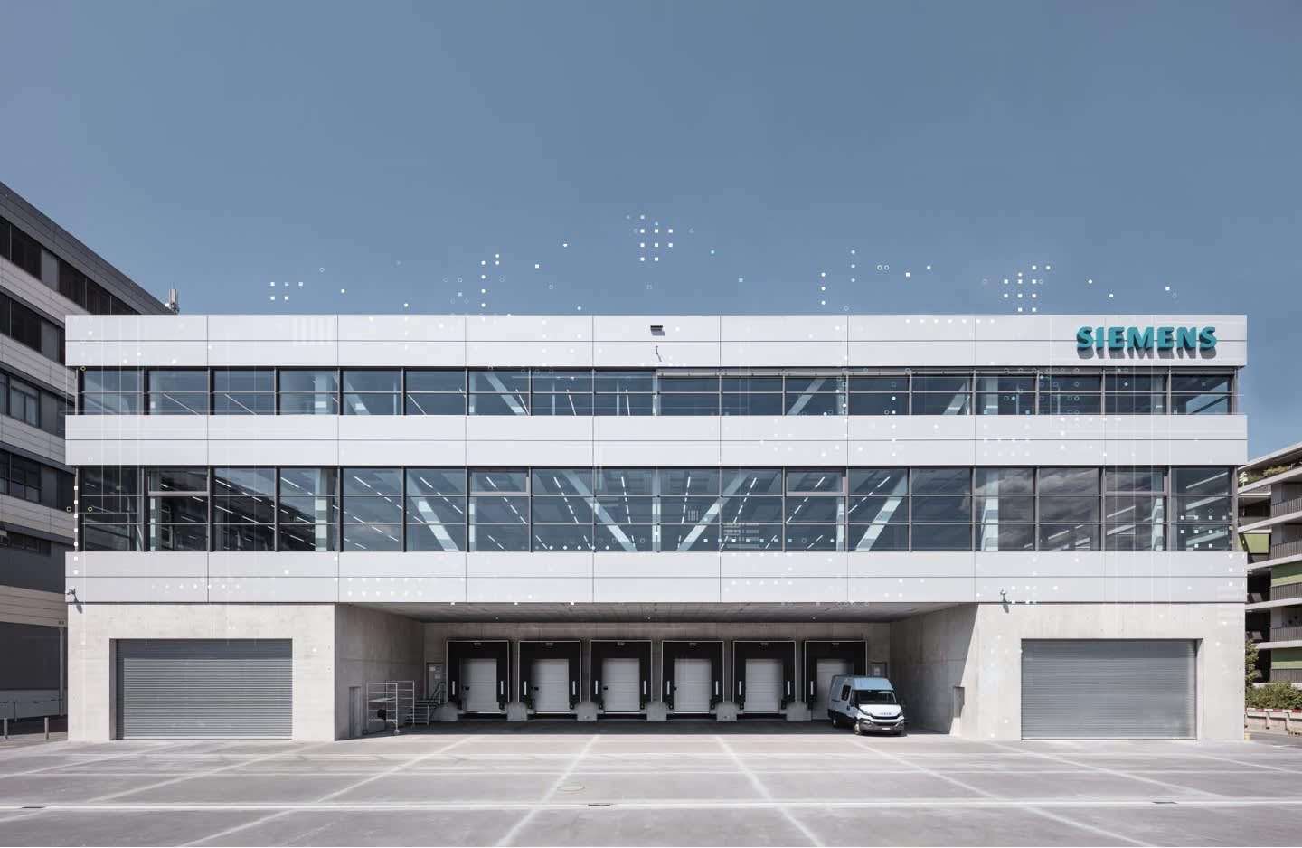 A modern three-story Siemens building with a glass facade, loading docks on the ground level, a parked white van, and the Siemens logo visible on the top right corner.