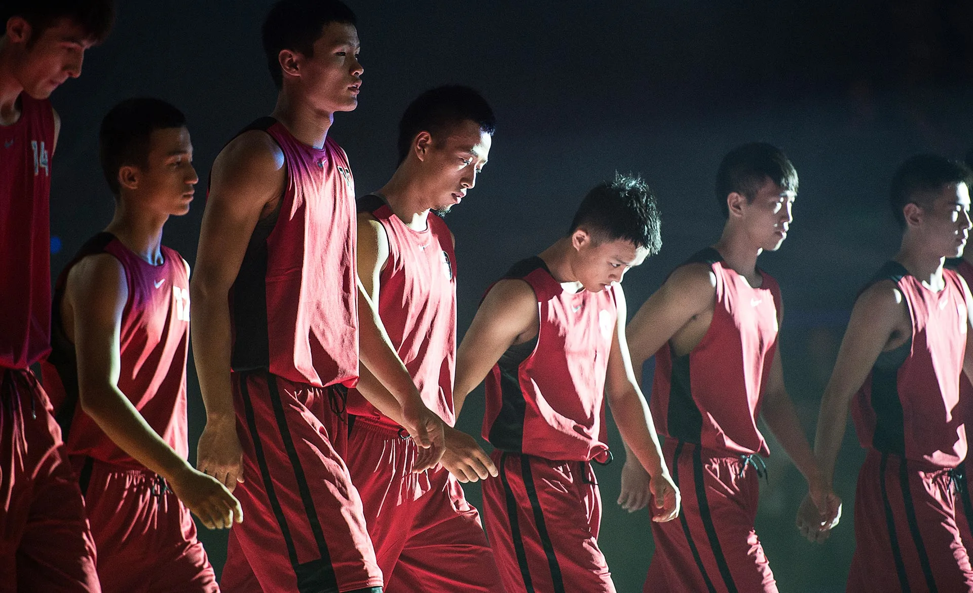 A group of young male basketball players in matching red uniforms walk together in a line under dramatic lighting, looking focused and serious.