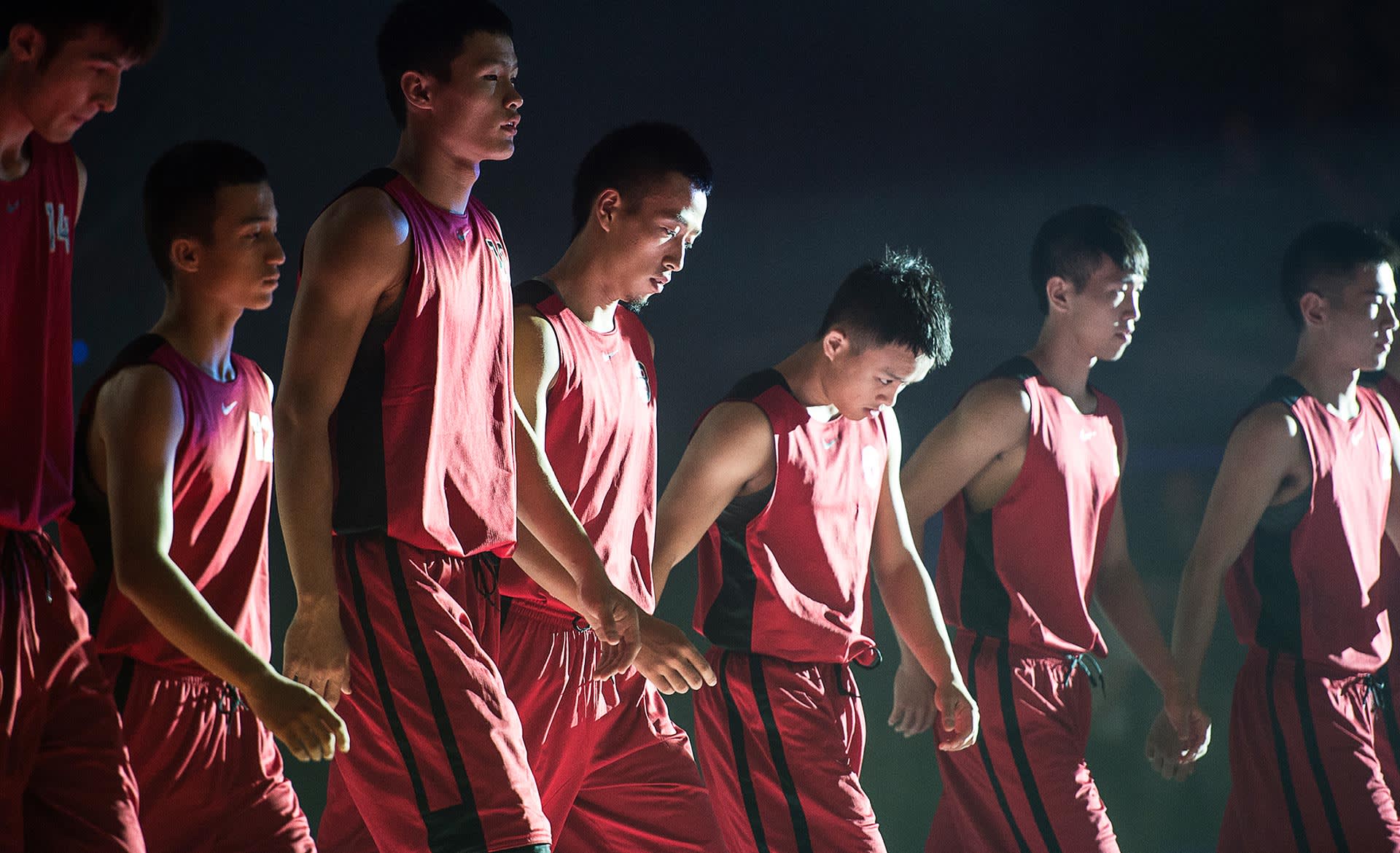 A group of young male basketball players in matching red uniforms walk together in a line under dramatic lighting, looking focused and serious.