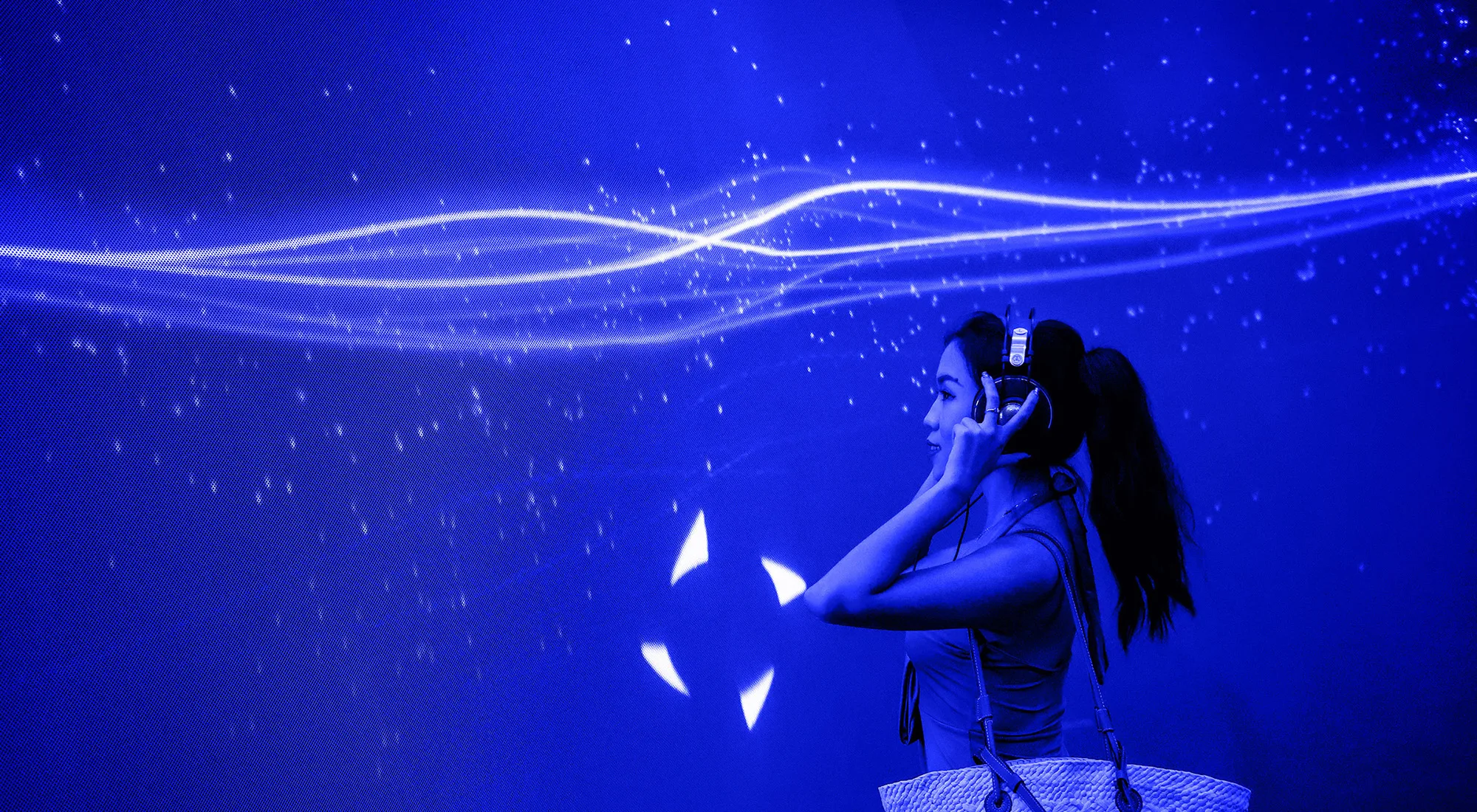 A woman wearing headphones stands against a glowing blue background with dynamic light trails and sparkles, seemingly immersed in a digital or interactive experience.