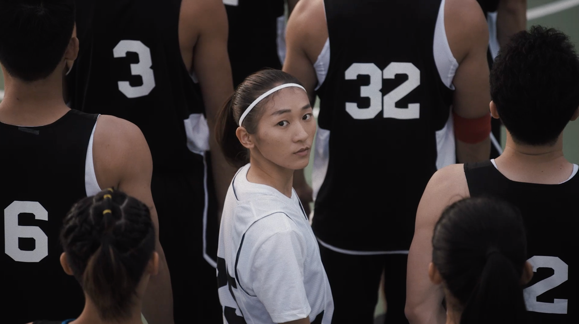 A person in a white shirt and headband stands among others in black basketball jerseys, looking back toward the camera while the rest face away. The numbers 3, 6, and 32 are visible on the jerseys.