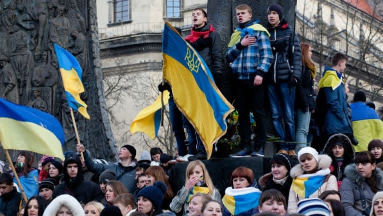 A large group of people gather outdoors, many holding or wearing Ukrainian flags. Some stand on a monument, one singing with hand on heart. The crowd appears peaceful and unified.