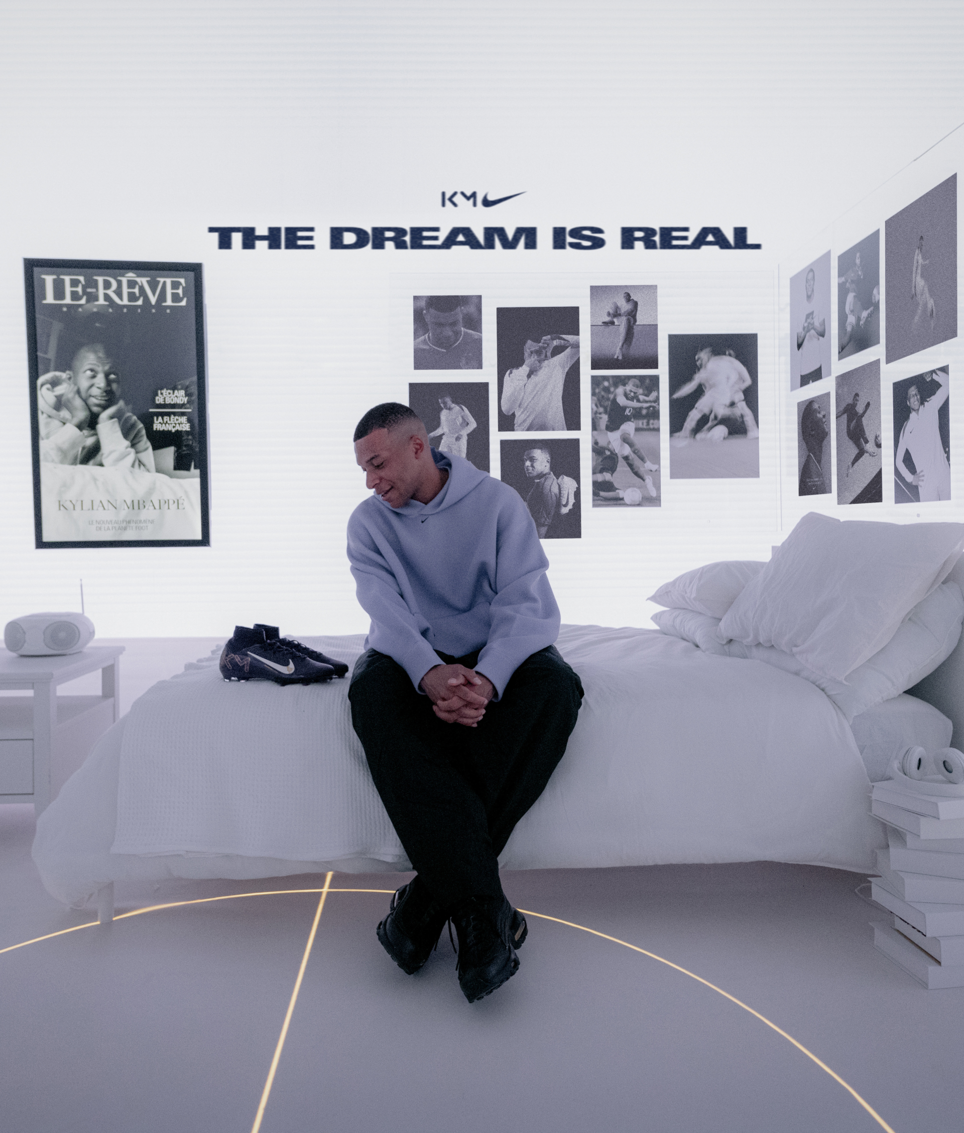 A young man sits on a white bed in a bright room with sports photos on the wall, a magazine cover, and THE DREAM IS REAL and a Nike logo above him. A pair of sneakers is on the bed next to him.
