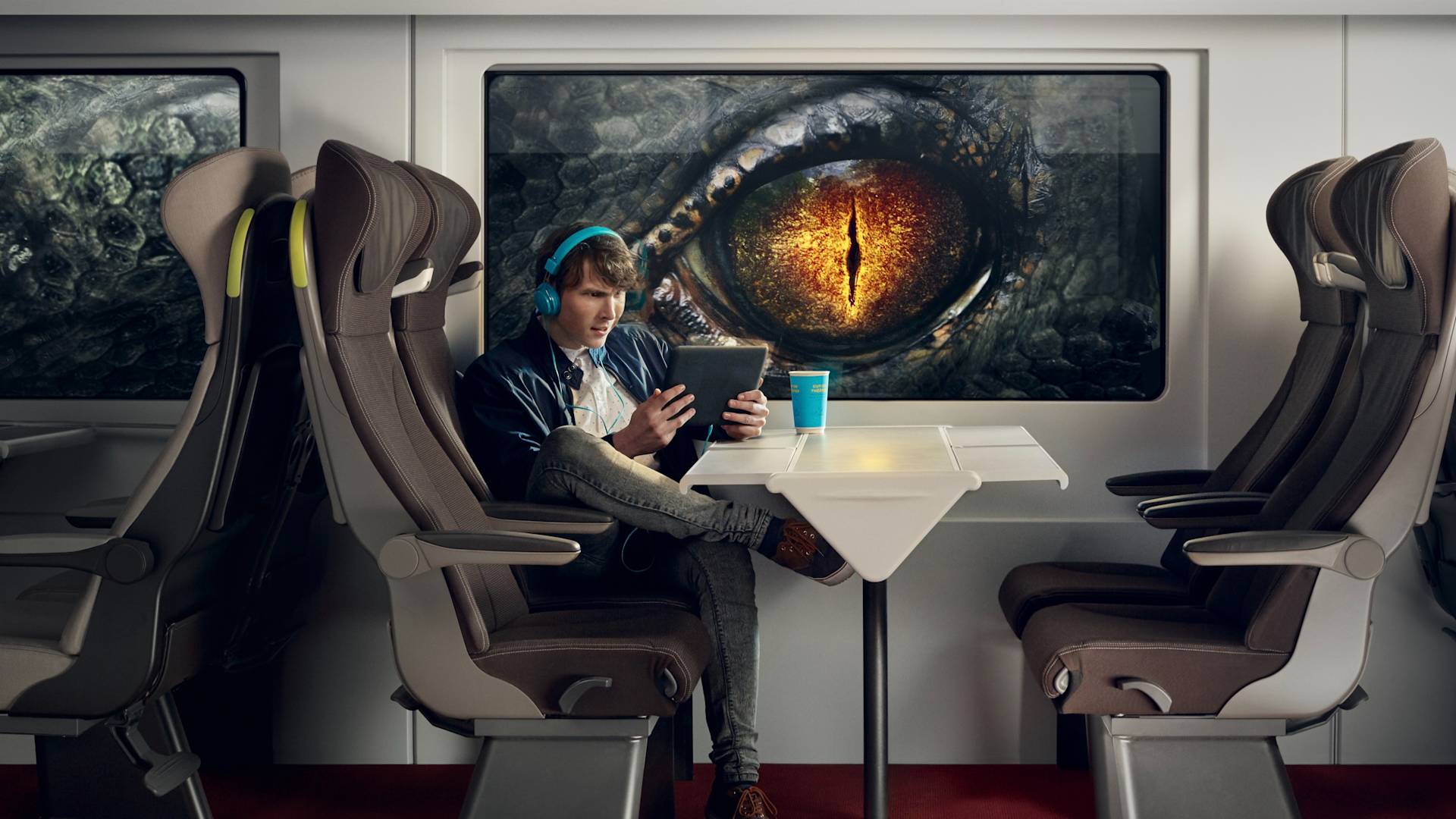 A person wearing headphones uses a tablet while sitting alone on a train. Outside the window, a giant reptilian eye peers in, filling most of the view. A blue travel mug sits on the table in front of the person.