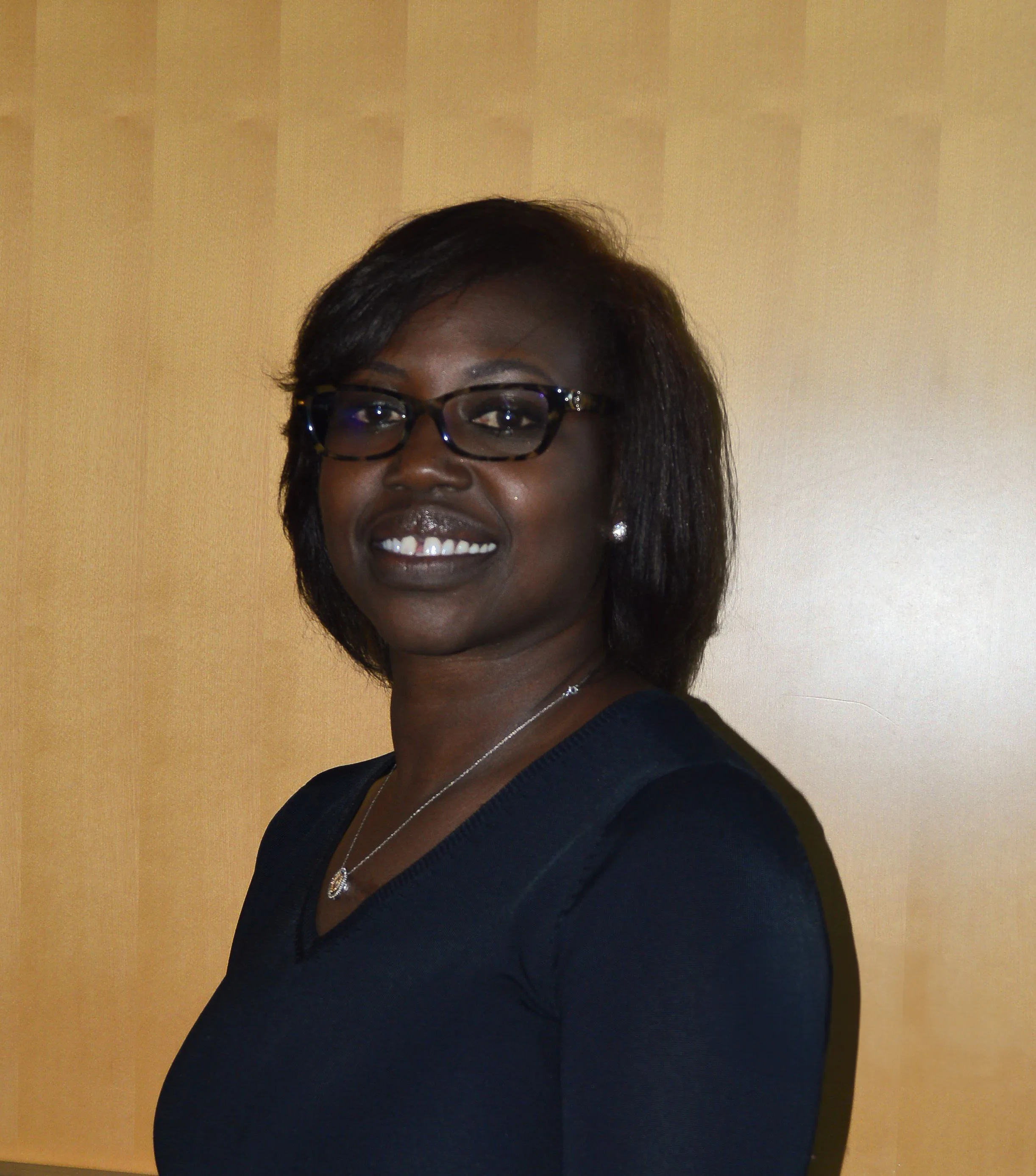 A woman with short dark hair, wearing glasses, a dark top, a necklace, and pearl earrings, stands smiling in front of a light wooden background.