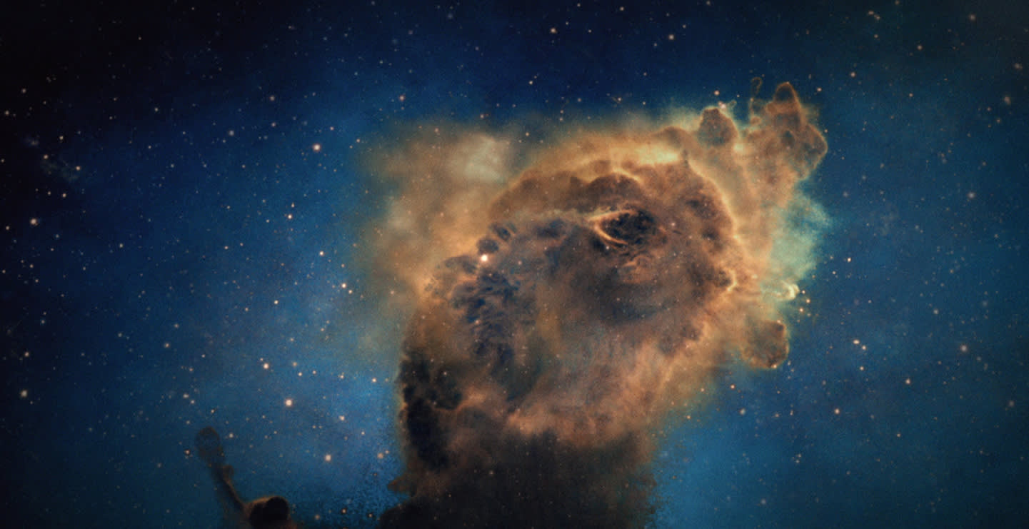 A colorful interstellar cloud of gas and dust glows against a dark, star-filled background in space, showing intricate shapes and bright blue and orange hues.