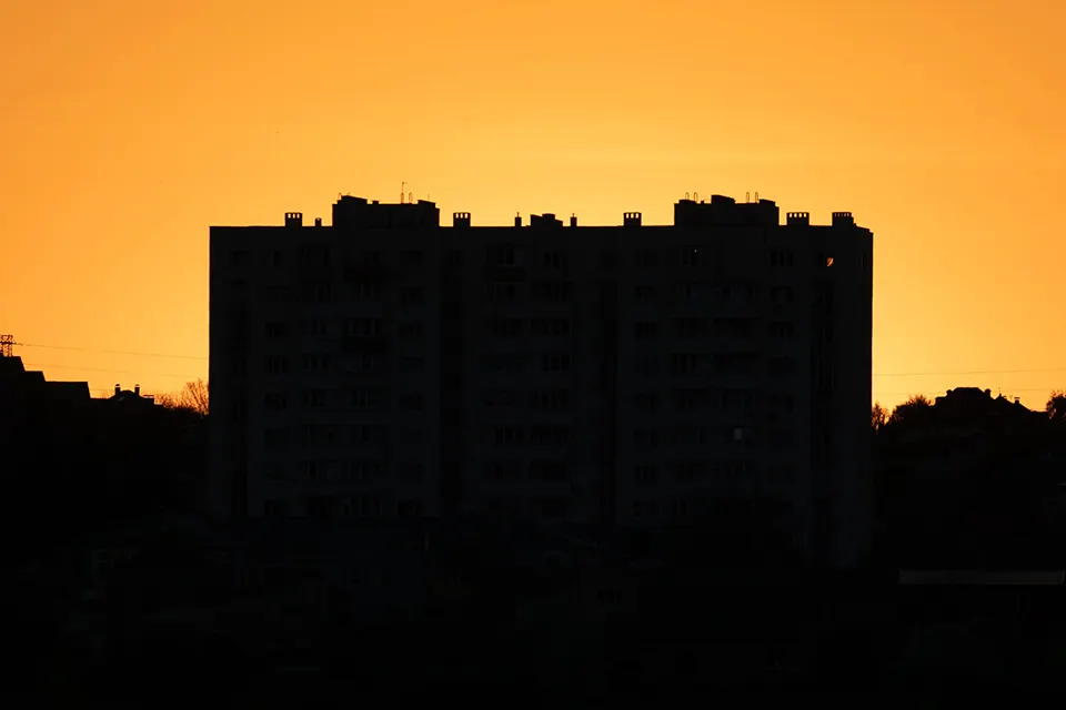 A tall apartment building is silhouetted against a bright orange sunset sky, with the structure appearing dark and featureless.