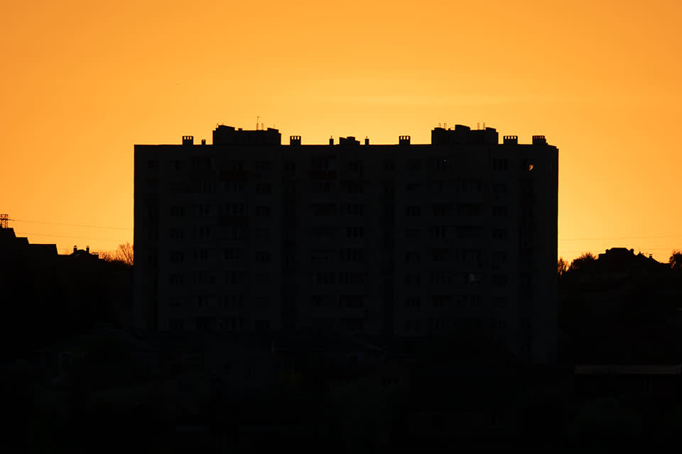 A tall apartment building is silhouetted against a bright orange sunset sky, with the structure appearing dark and featureless.